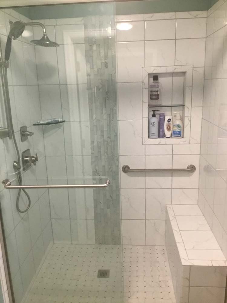 Shower with glass door, white tiled walls, built-in bench, grab bar, and recessed shelf with toiletries.