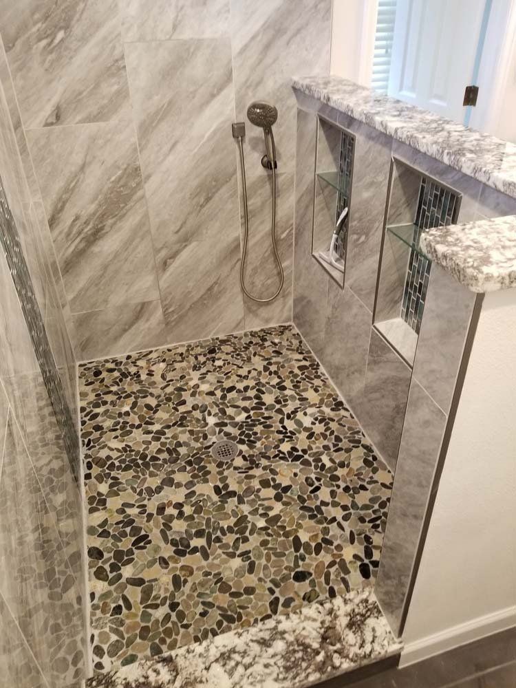Walk-in shower with pebble floor, gray tile walls, and built-in shelves. Granite countertop edges.
