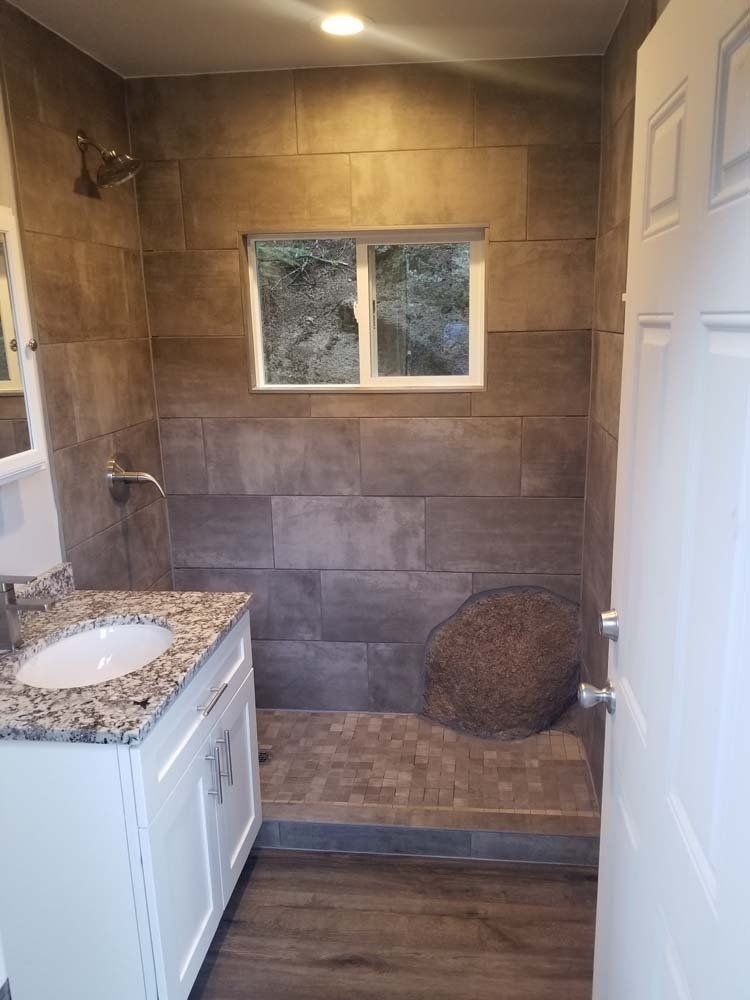 Bathroom with brown tiled shower and walls, white vanity, window with outdoor view, and open door.