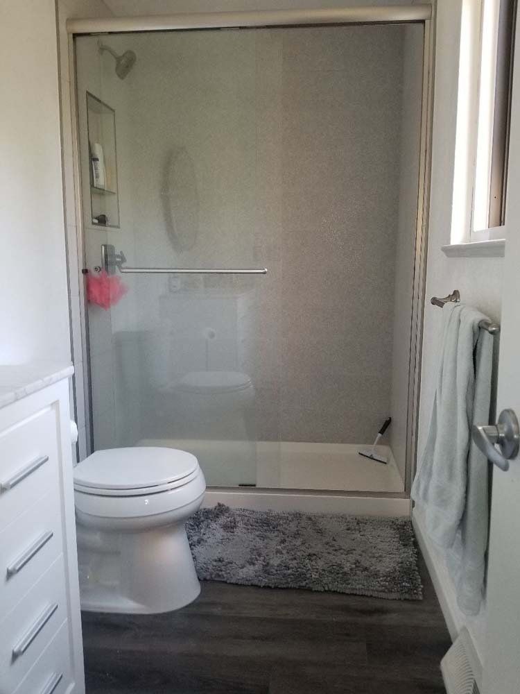 Bathroom with a glass-enclosed shower, toilet, and vanity. Gray rug and towel. White walls and fixtures.