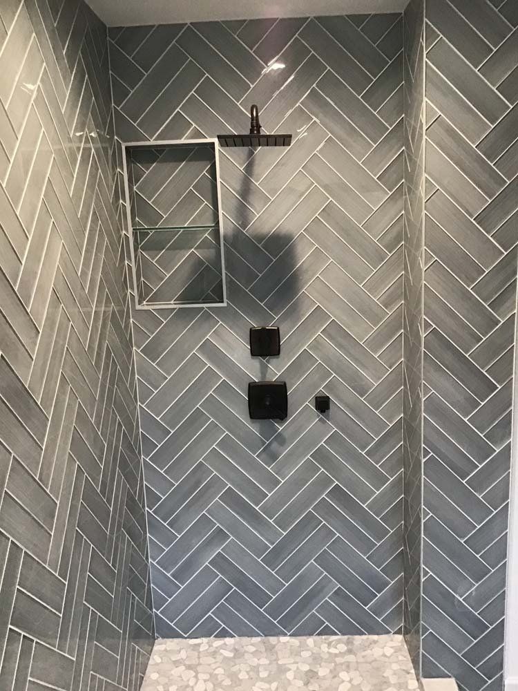 Grey tiled shower with herringbone pattern. Black shower head and fixtures. Glass shelf on the left.