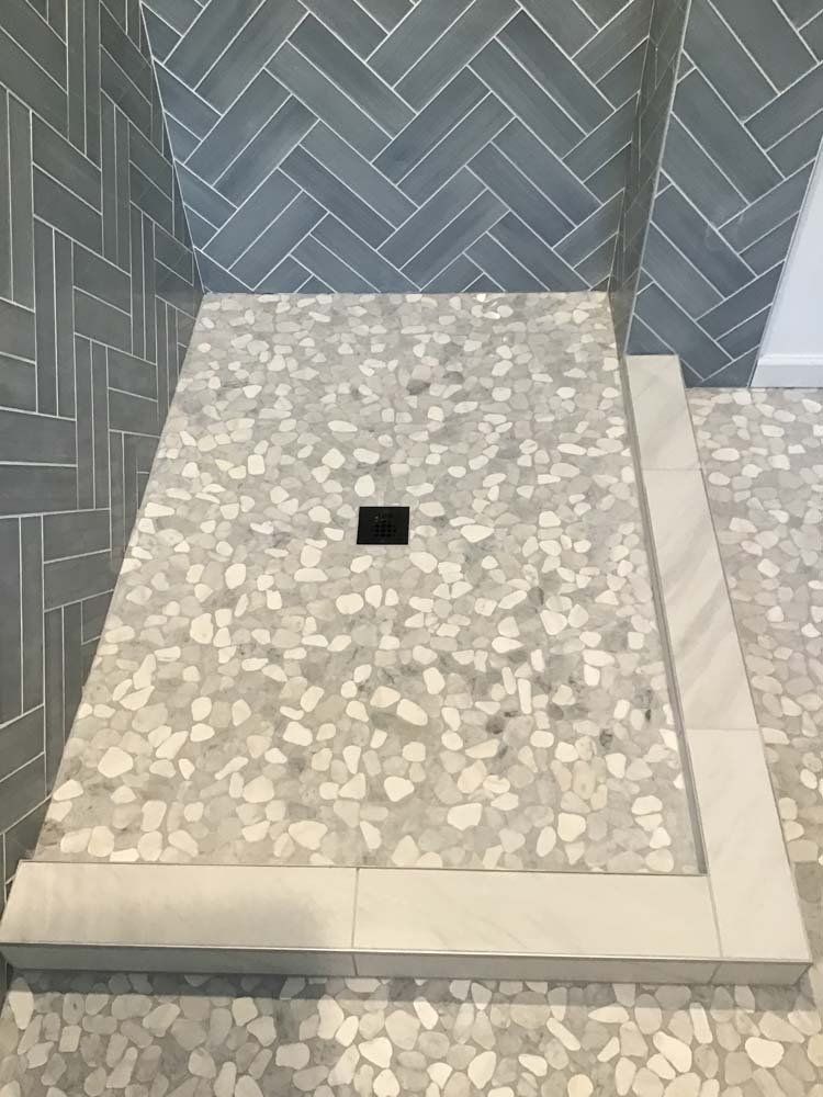 Shower with gray pebble floor and herringbone-patterned wall tiles. White curb surrounds.