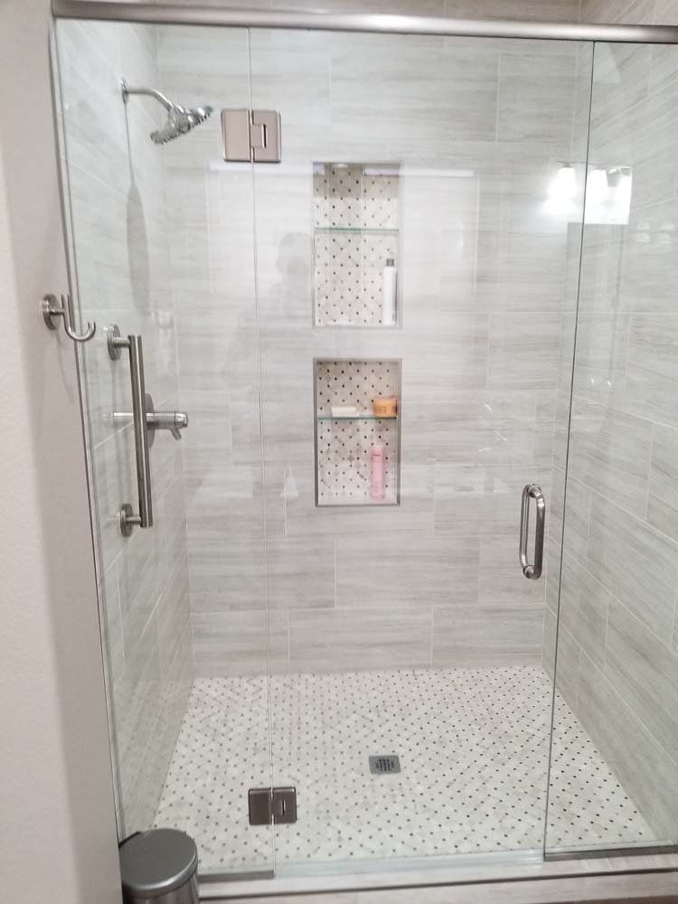 Glass-doored shower with gray and white tile, two built-in shelves, and a silver shower head and door handle.