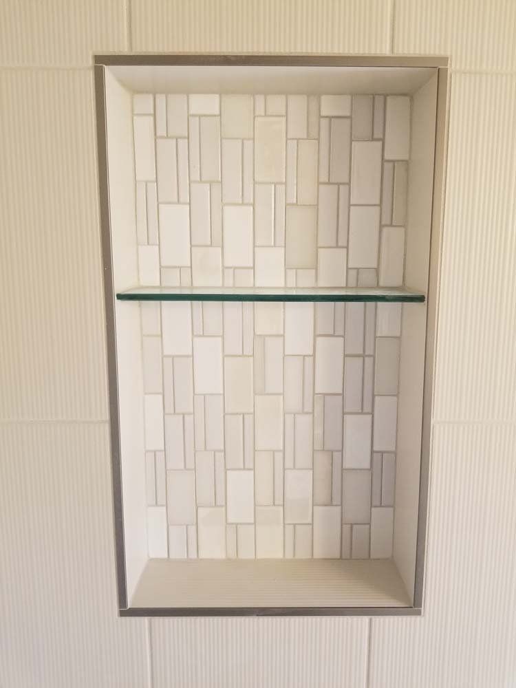 Built-in bathroom shower niche with glass shelf and mosaic tile backing, surrounded by light colored tiles.