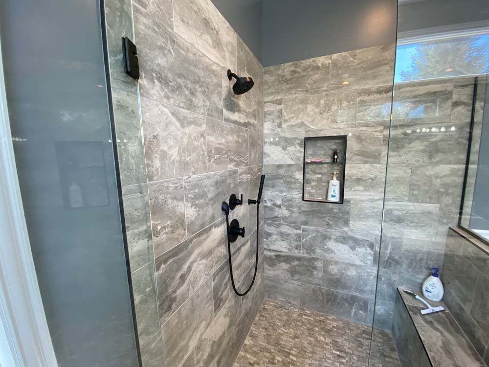 Modern shower with gray tiled walls, black fixtures, and a glass door. Pebble floor and a built-in shelf.