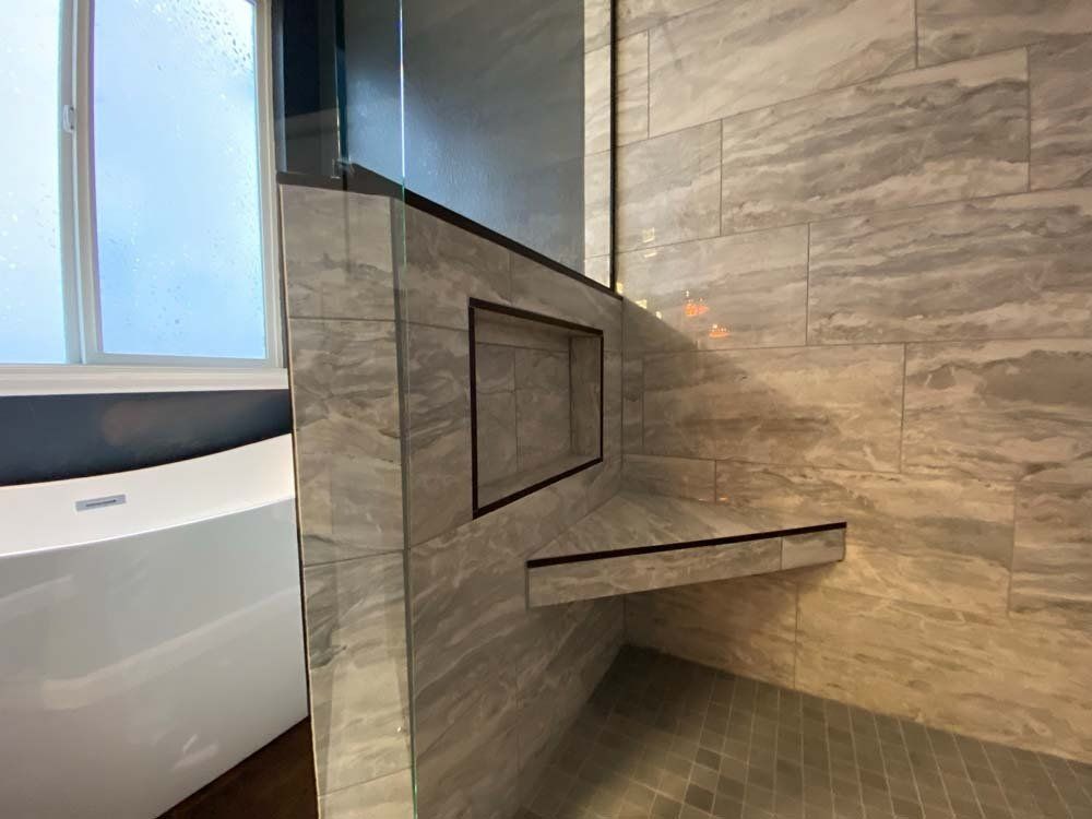 Modern bathroom with gray tiled shower and built-in shelf and niche. Glass shower door and white tub visible.