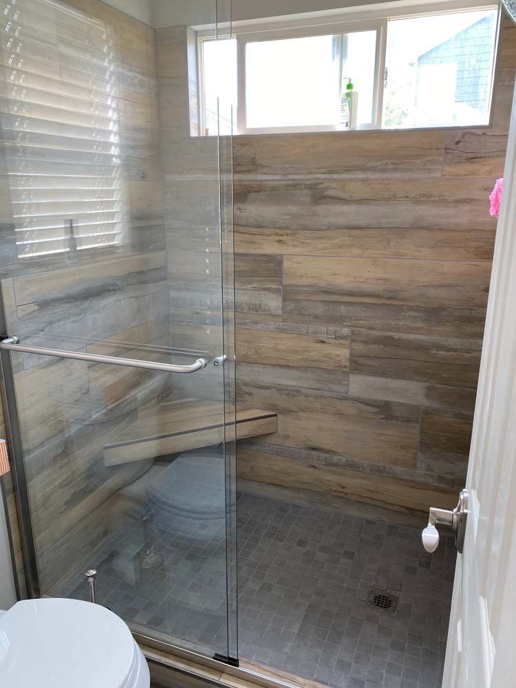 Shower with wood-look tile walls, glass door, and small window. A toilet sits next to the shower.