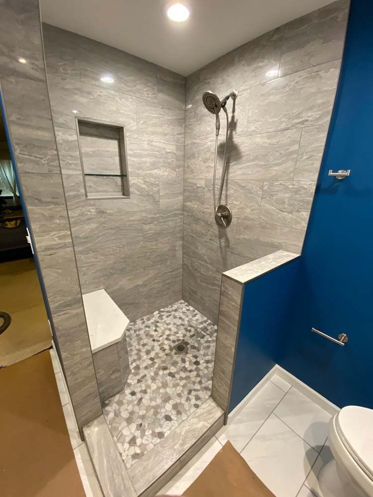 Walk-in shower with grey tile walls, pebble floor, built-in seat, and blue accent wall.