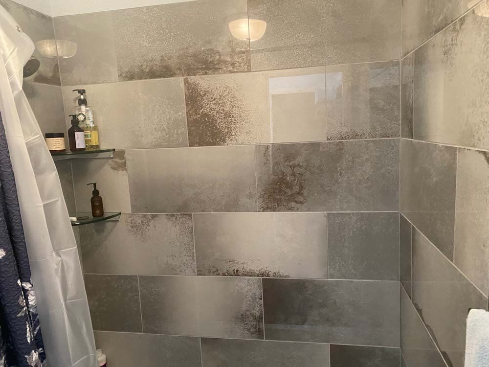 Grey tiled shower with glass shelves holding bottles and a light fixture.