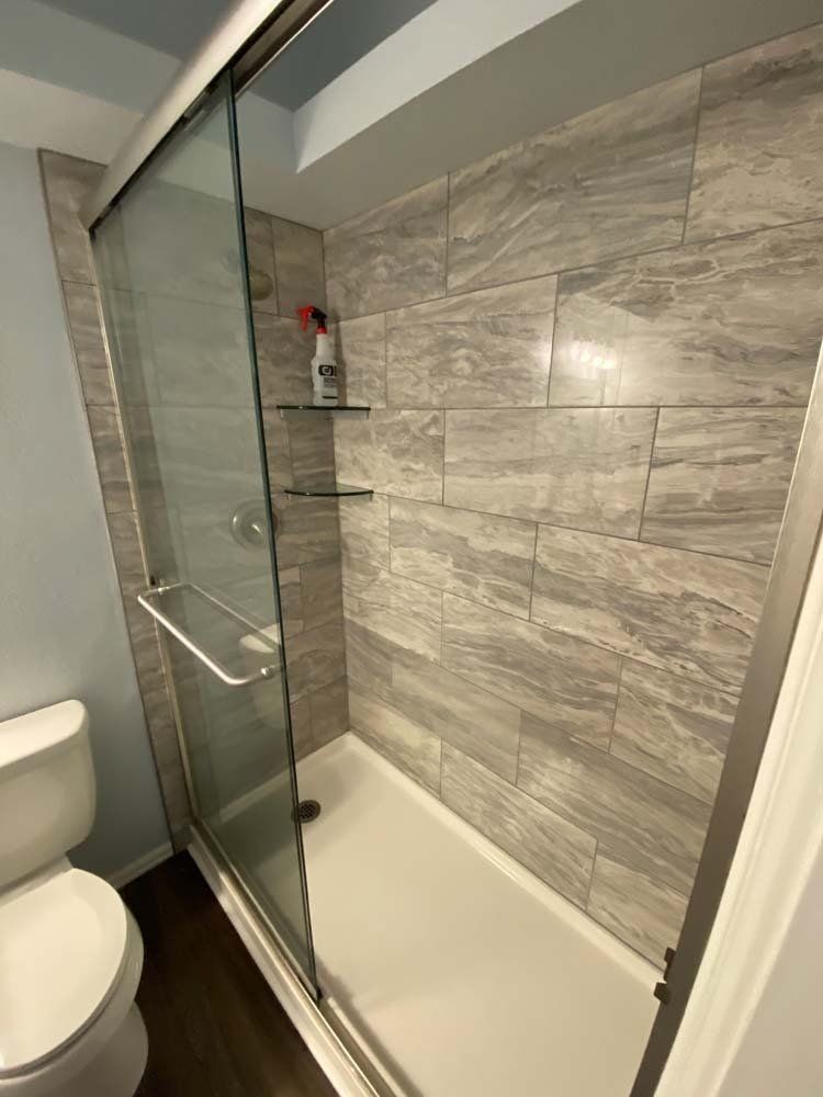 Shower with sliding glass doors, gray tiled walls, two glass shelves, and a toilet.
