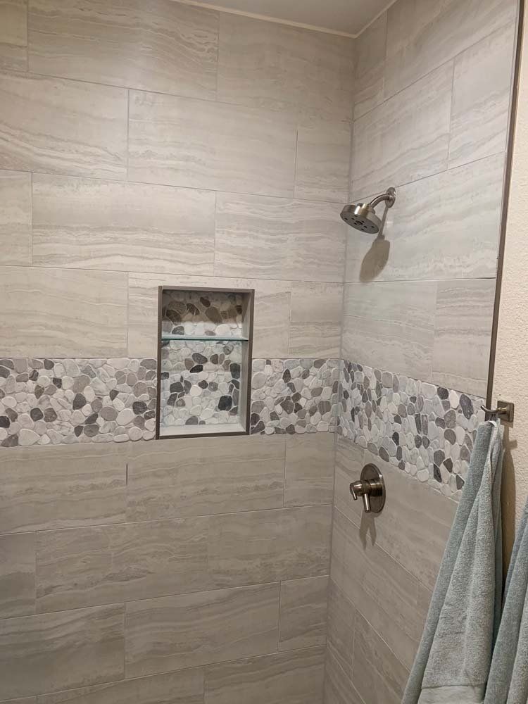 Shower interior with light gray tiles and mosaic border. Includes a built-in shelf and showerhead.