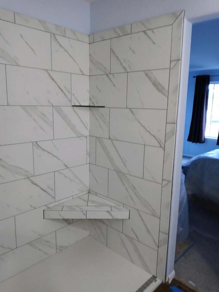 Newly tiled shower stall with corner shelf and seat; white marble-look tiles.