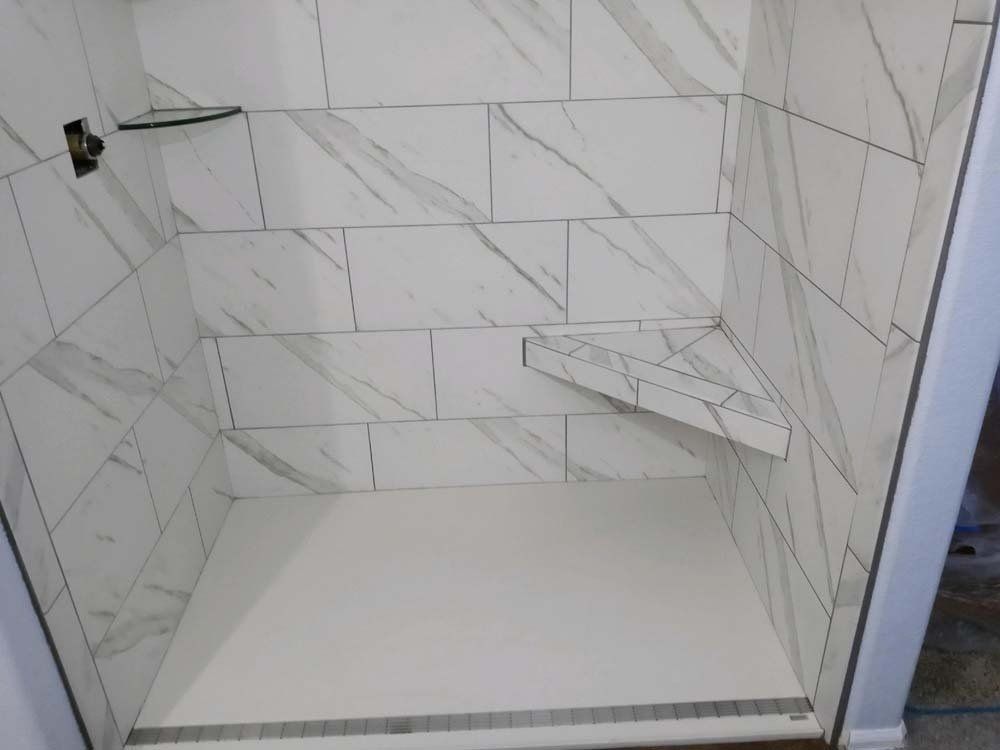 Shower stall with white marble-look tiles, a corner shelf, and a built-in bench.