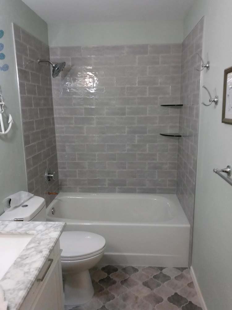 Bathroom with gray tiled shower, white tub and toilet, marble vanity, and patterned floor.