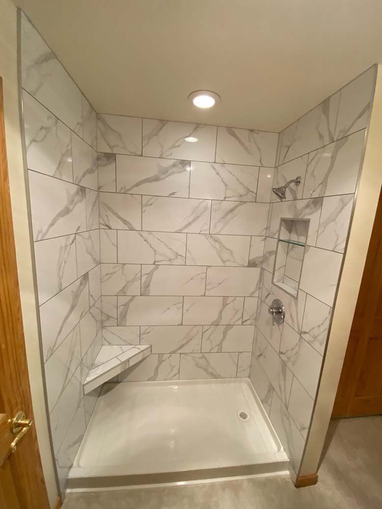 Newly tiled shower with white marble-patterned tiles, showerhead, built-in shelf, and corner seat.