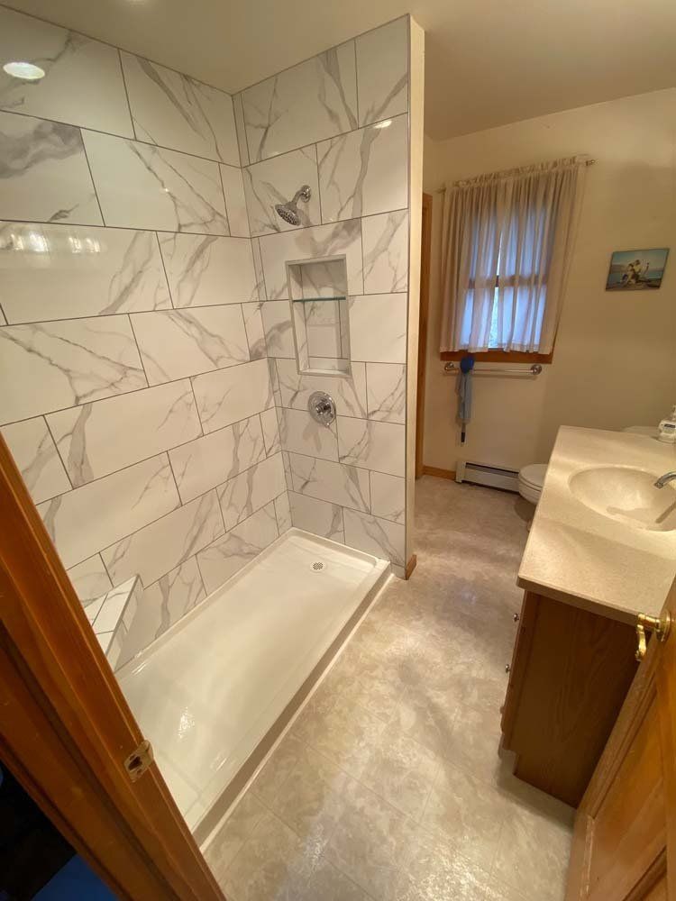 Newly renovated shower stall with white marble-look tile, next to a vanity in a bathroom with beige walls.