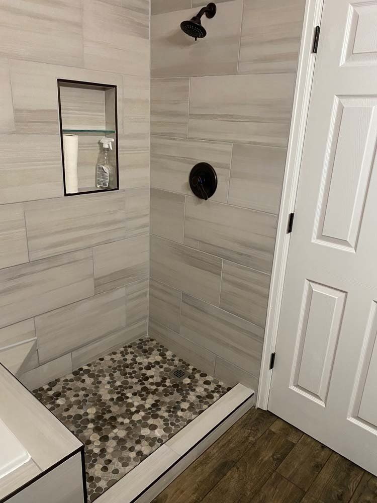 Shower with light gray tiled walls and pebble floor. Built-in shelf, black shower fixtures, white door.