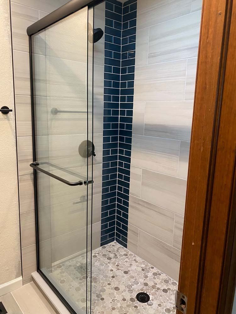 Shower stall with glass door, black fixtures, blue tile accent, pebble floor.