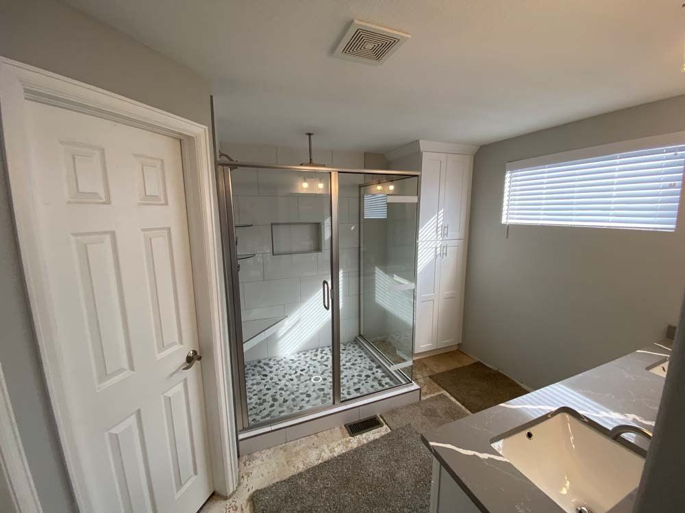 Bathroom with glass shower, white door, cabinets, gray walls, and window with blinds.