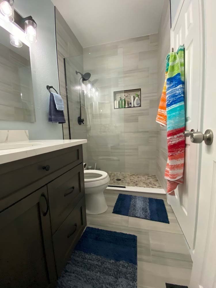 Bathroom with gray tile shower, dark vanity, blue rugs, and towels hanging on the door.