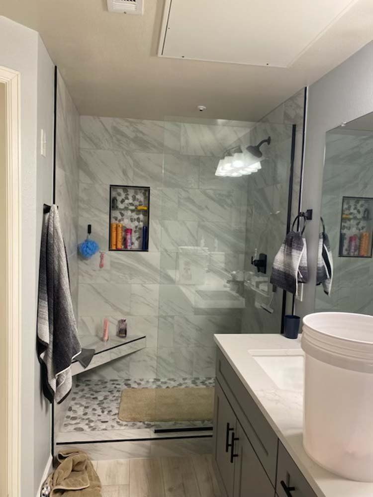 Modern bathroom with glass shower, gray vanity, and marble-look tile. Towels hang on black bars.
