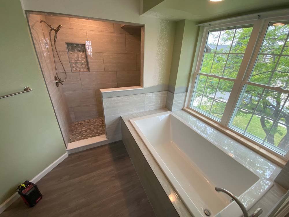 Bathroom with shower and tub; light-colored tiles, green walls, large window with trees outside.