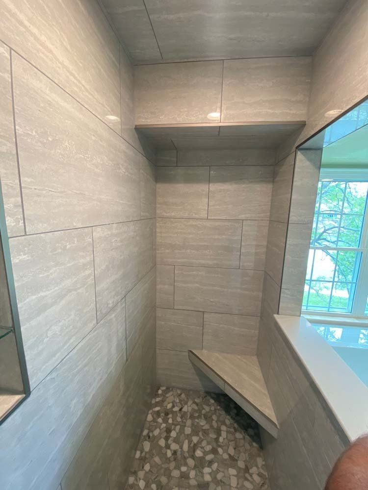 Tiled shower with built-in bench and shelf. Pebble floor, gray tile walls. Bright window to the right.