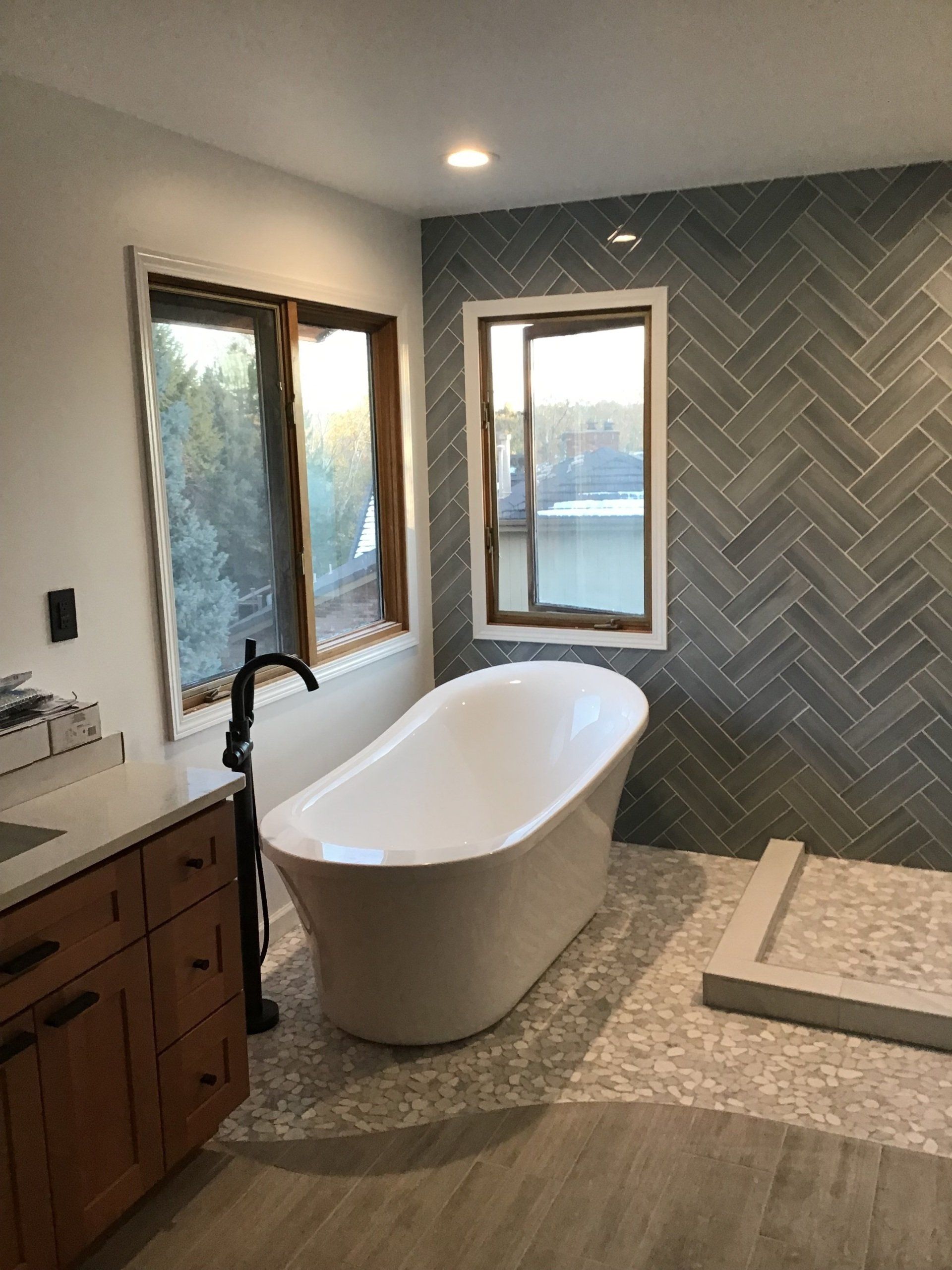 Modern bathroom with a white freestanding tub, wood windows, and gray herringbone tile.