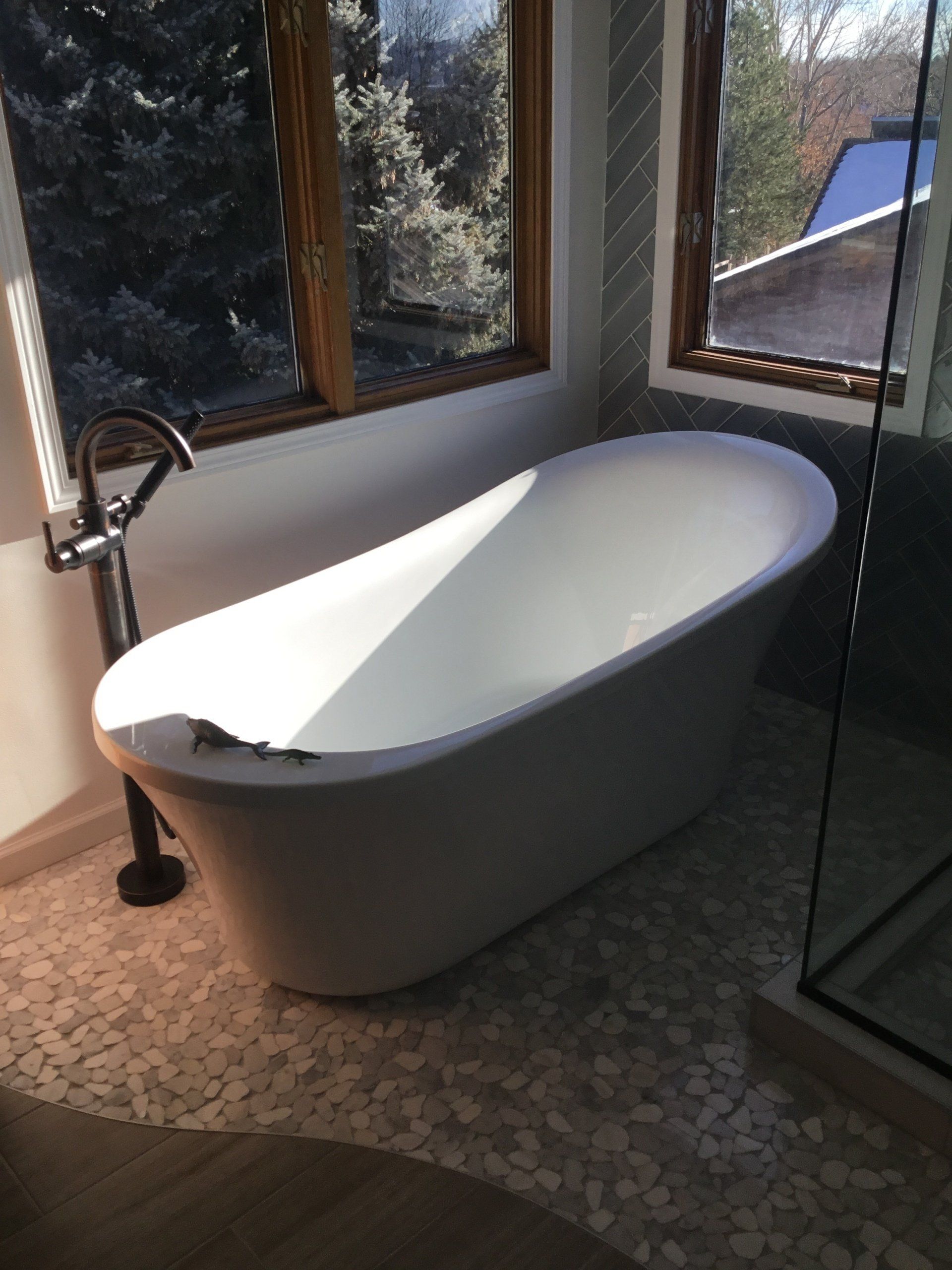 Freestanding white bathtub by a window in a bathroom with patterned tile flooring.