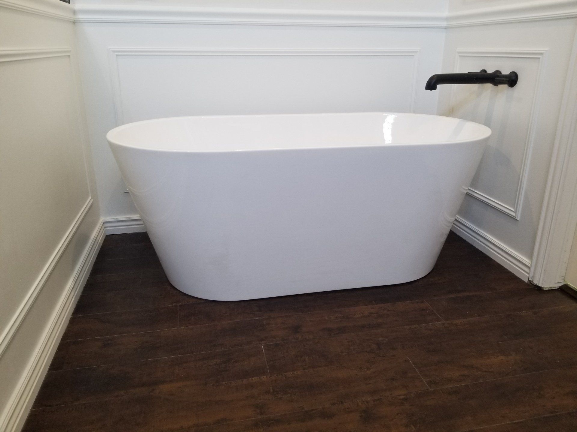 White oval bathtub in a corner with dark wood floor and black faucet.