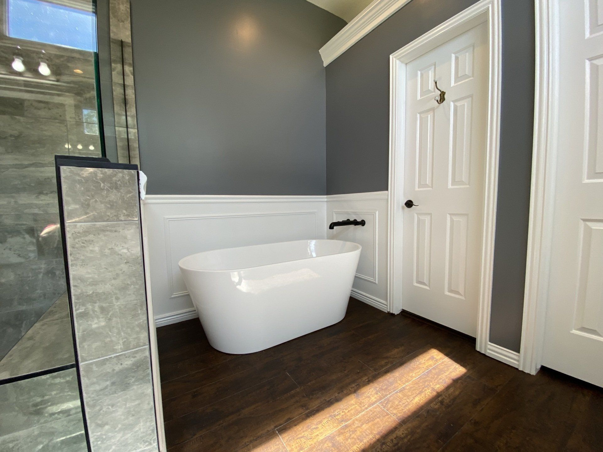 Modern bathroom with a white freestanding tub against a gray wall, dark wood floor, and a white door.
