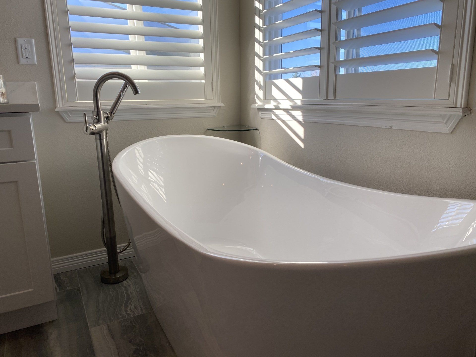 Freestanding white bathtub in a bathroom with window shutters and a tall faucet.