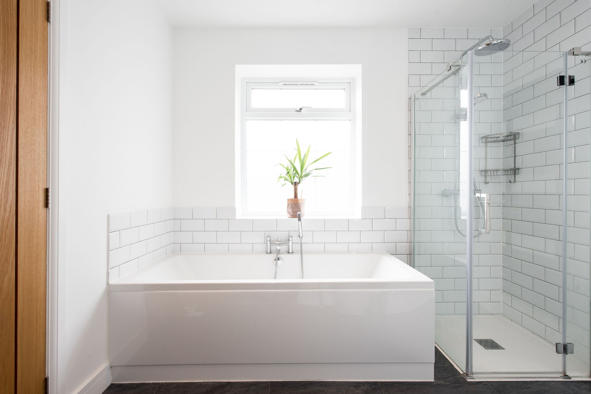 White bathroom with tub, shower, and window; white brick-like tile, dark floor, and wooden door.