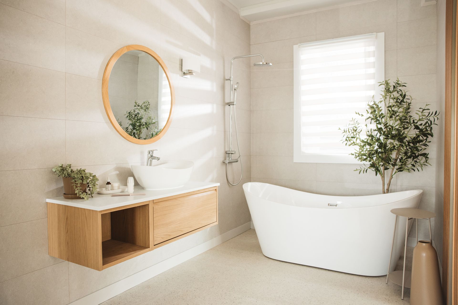 Modern bathroom with a white tub, wooden vanity, and oval mirror. A window lets in sunlight.