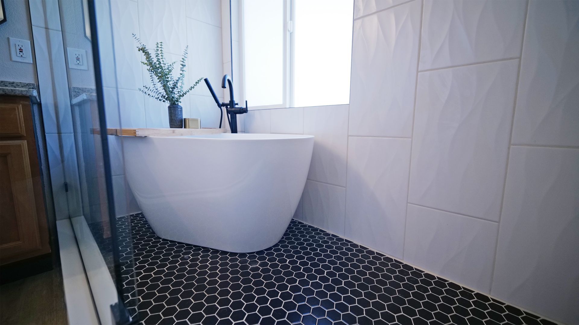 Modern bathroom with white freestanding tub, black hexagon tile floor, and white textured wall.