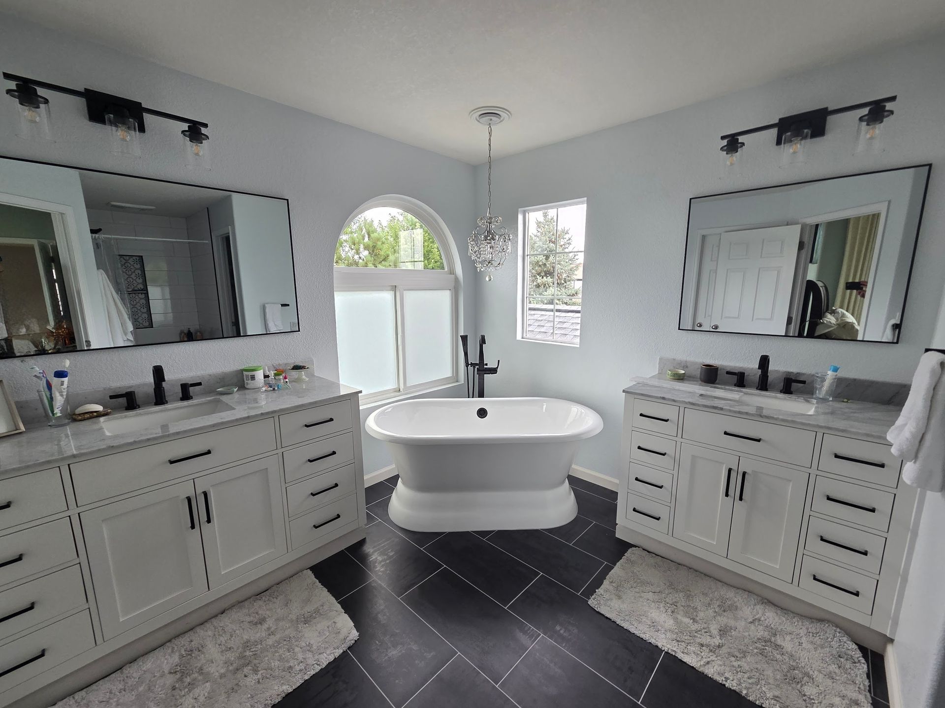 Bathroom with dual sinks, a freestanding tub, and a window. White cabinets, black fixtures, and dark tile floor.