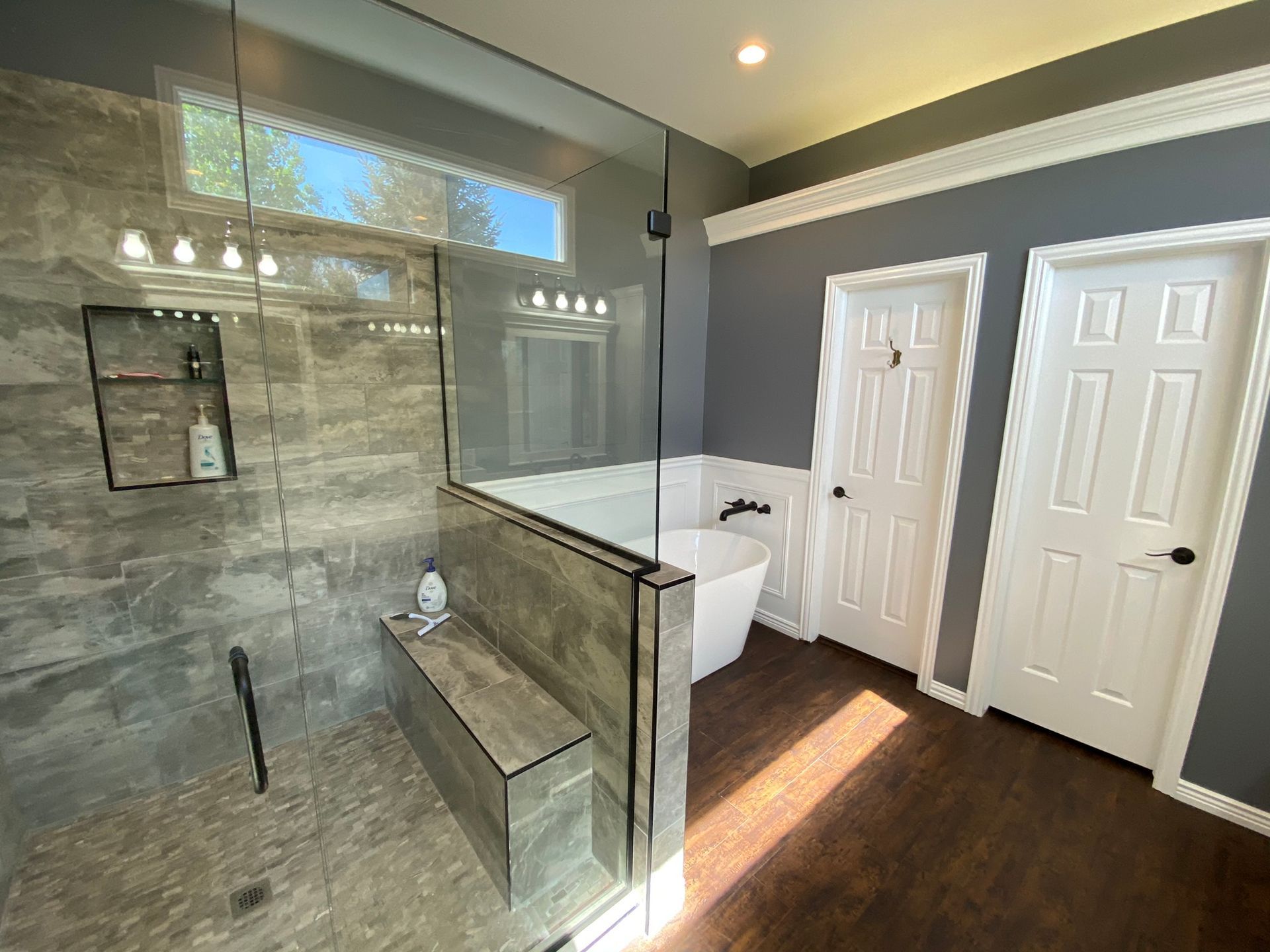 Modern bathroom with gray tile shower, glass door, white tub, two white doors, dark wood floor.