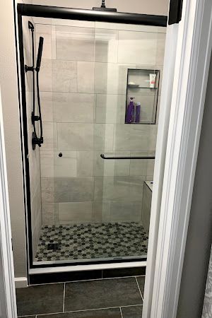 Black-framed glass shower with pebble floor, gray tiled walls, and built-in shelf.