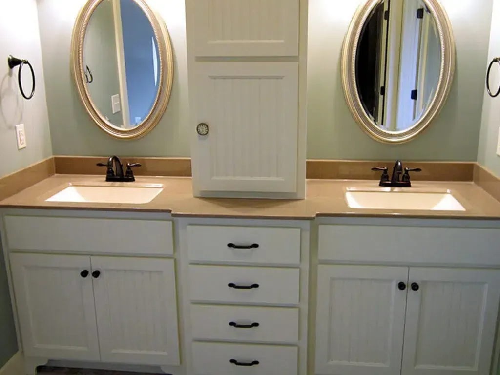 Bathroom with dual sinks, cream cabinets, and tan countertops. Oval mirrors, dark faucets, and light green walls.
