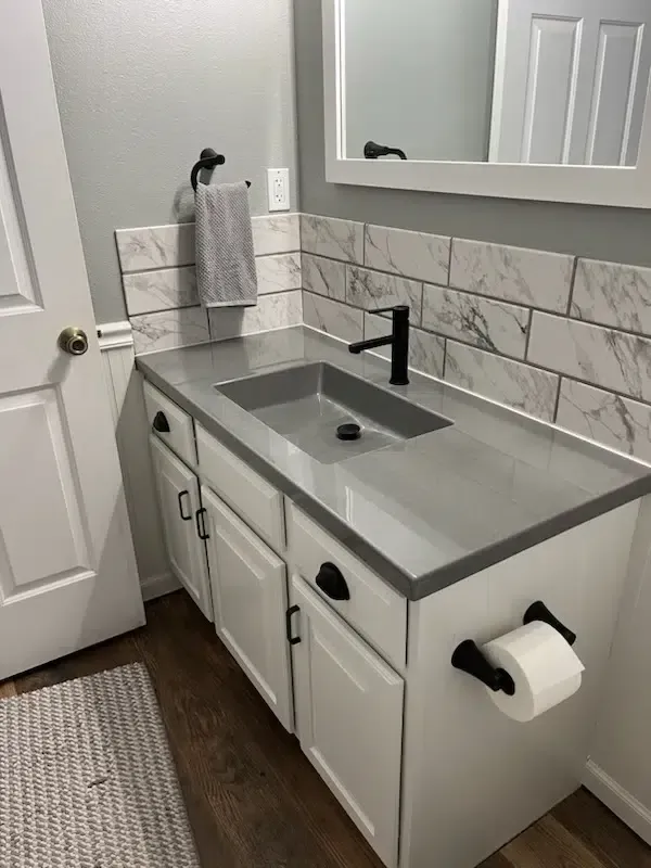 Bathroom vanity with gray countertop, white cabinets, black faucet, and marble tile backsplash.