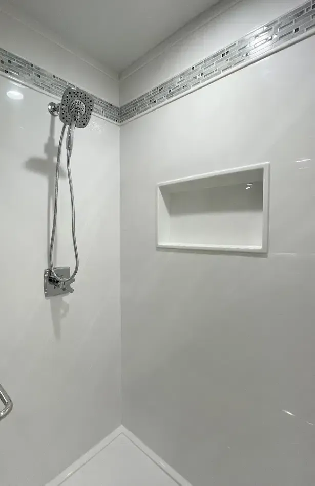 White-walled shower with corner entry, built-in shelf, and silver shower head and fixtures. Decorative tile border.