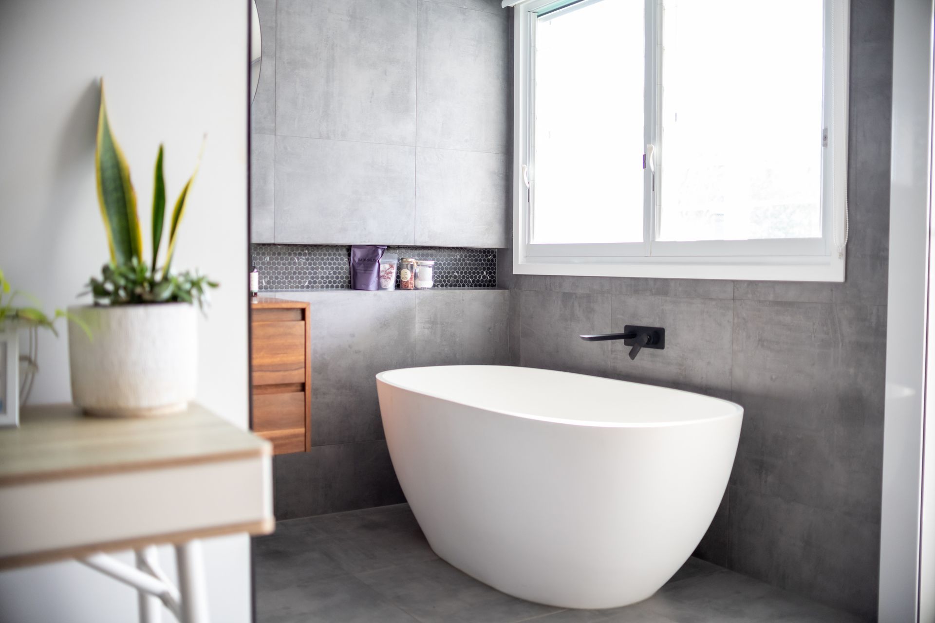 Modern bathroom with a white oval bathtub, gray tiled walls, and a small wooden cabinet.