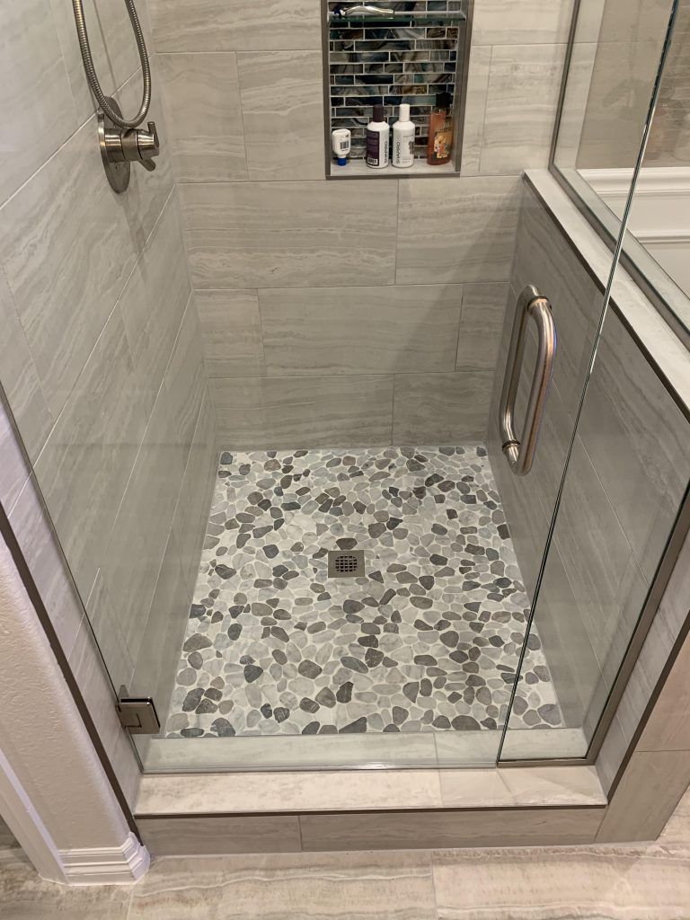 Bathroom shower with glass door. Gray tile walls and floor with pebble-like tiles. Recessed shelf.