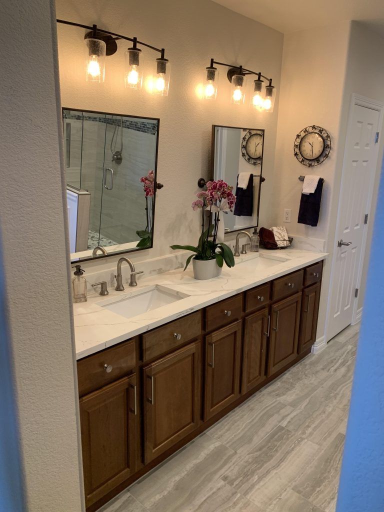 Bathroom with dual sinks, mirrors, brown cabinets, and light fixtures. A plant sits on the countertop.