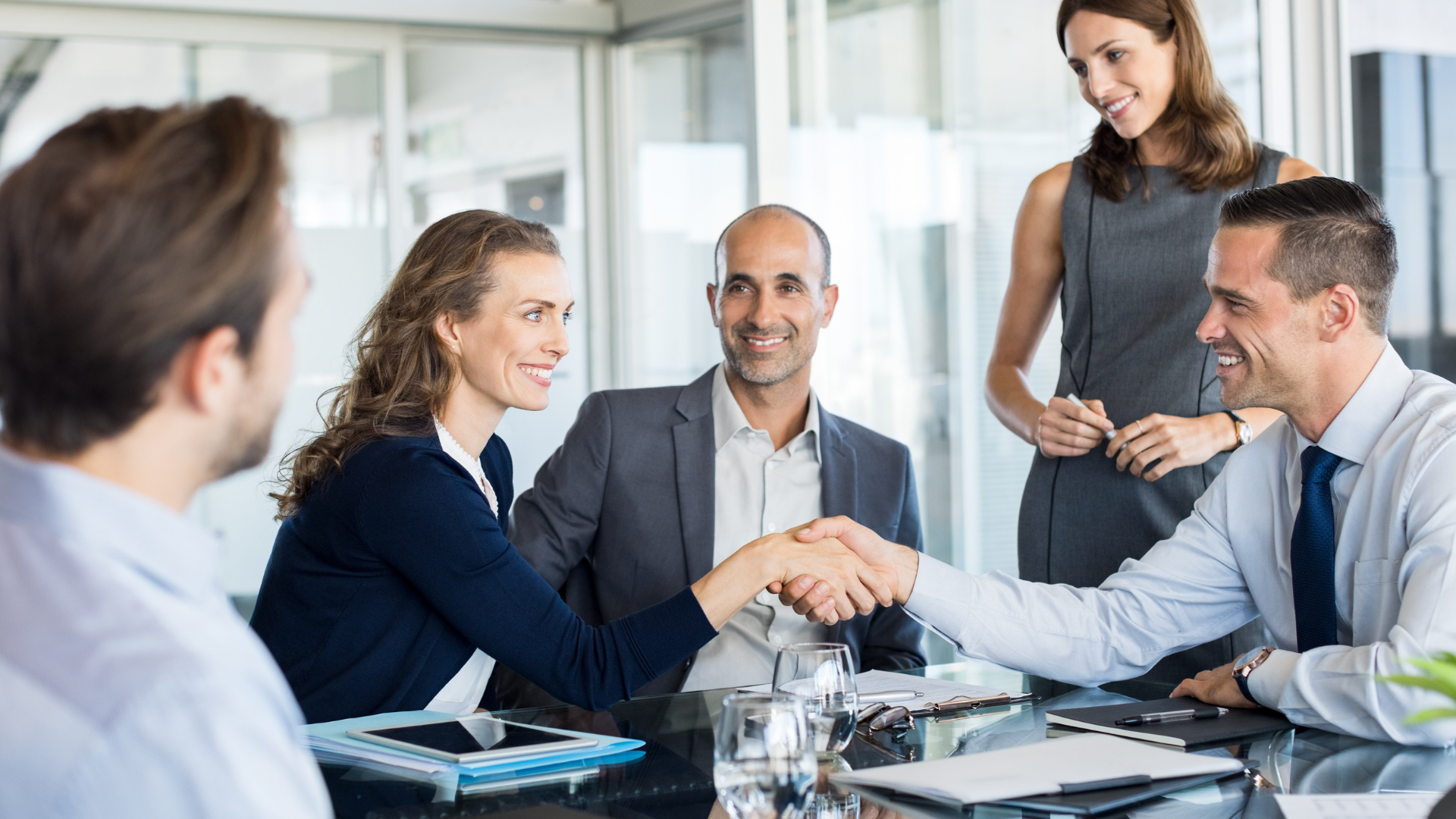 Business colleagues in a meeting shaking hands across a conference table, smiling in an office.
