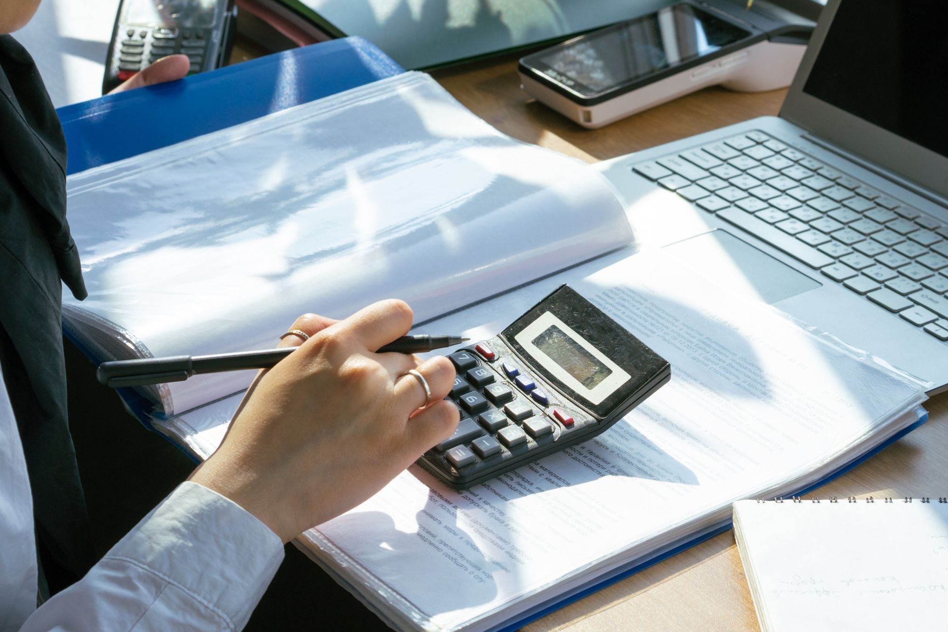 Person using a calculator beside an open laptop and blue binder on a desk