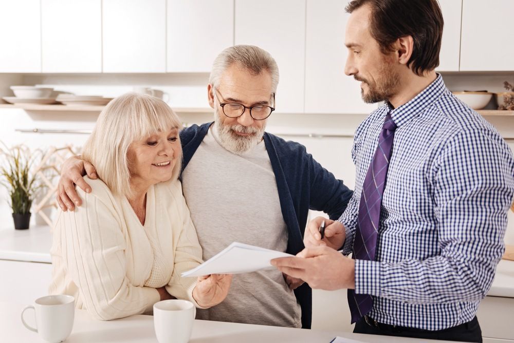 A Man is Talking to an Elderly Couple in a Kitchen While Holding a Piece of Paper — McVittie Legal In Ballina, NSW