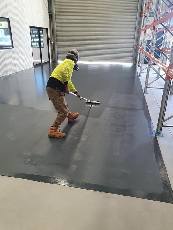 A Man Is Painting A Warehouse Floor With A Roller — LMS Painting & Maintenance Pty Ltd In North Lambton, NSW