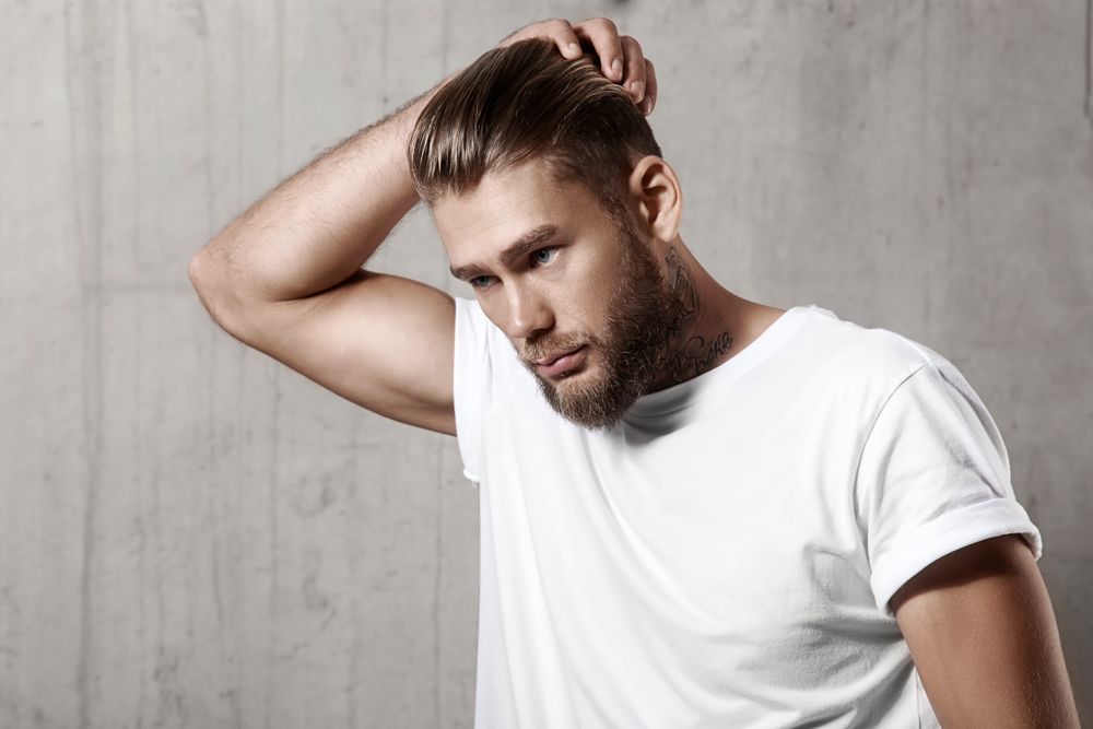 A man with a beard is wearing a white t-shirt and touching his hair.