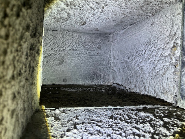 Interior of a rectangular duct filled with white, fuzzy material and dark debris at the bottom.